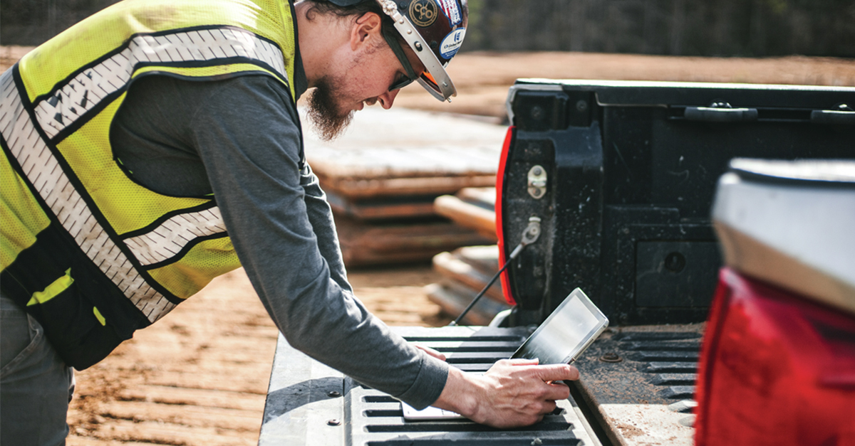 Construction worker looking at a laptop in the field on the back of a truck reviewing plans in Fieldwire on his tablet.